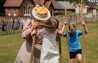 A girl and a boy on stilts being helped by a lady in Victorian costume.