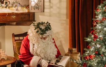 Father Christmas, wearing a red suit with white fur trim and a wreath of greenery on the head, sits in an armchair reading a book. The indoor scene fe
