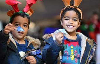 two young children wearing reindeer antlers and Christmas jumpers under their coats.