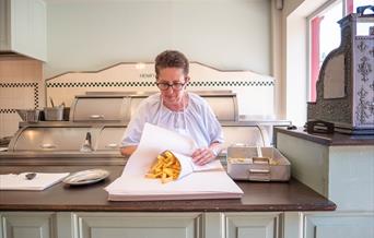 A lady wrapping traditionally cooked chips in paper in a Victorian chip shop at Blists HIll Victorian Town