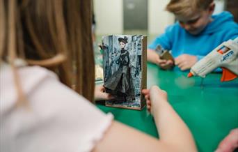 Girl playing with a mechanical toy that she has made to look like a Victorian doll, there is a boy in the background and an adult holding a glue gun t