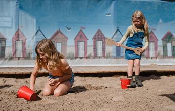 Two girls are playing in the sand with buckets and spades