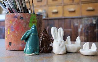 A picture taken in the studio, in the foreground a clay pot, covered in paint with brushes in the top and a selection of bunny figures in a row.