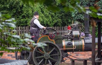 A man is seen standing on the back of a steam engine, driving it down the road at Blists Hill Victorian Town