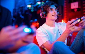 Teenage boy smiling and playing on a game, holding a controller, sat next to his friend.
