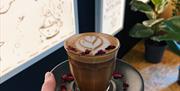 A photo of a hand holding out a drink in a glass on a saucer. The chocolatey drink has a pretty latte art patter with a heart, and a sprinkling of ros