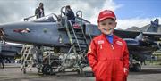 Little boy in red arrows overalls stood in front of an old plane