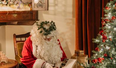 Father Christmas, wearing a red suit with white fur trim and a wreath of greenery on the head, sits in an armchair reading a book. The indoor scene fe