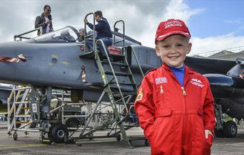 Little boy in red arrows overalls stood in front of an old plane
