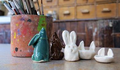 A picture taken in the studio, in the foreground a clay pot, covered in paint with brushes in the top and a selection of bunny figures in a row.