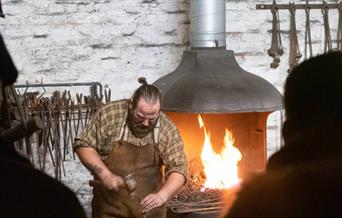 The image shows a blacksmith wearing a leather apron, in front of the forge.