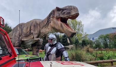 The Mandalorian standing by a vehicle with a Hoo Zoo T-Rex behind.
