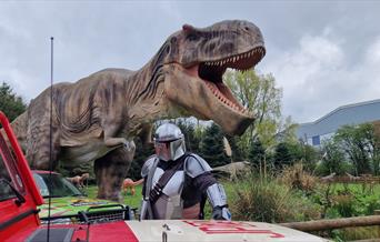 The Mandalorian standing by a vehicle with a Hoo Zoo T-Rex behind.