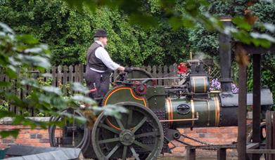 A man is seen standing on the back of a steam engine, driving it down the road at Blists Hill Victorian Town
