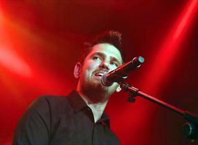 Scouting For Girls lead singer Roy Stride on stage in front of a red background singing into a microphone