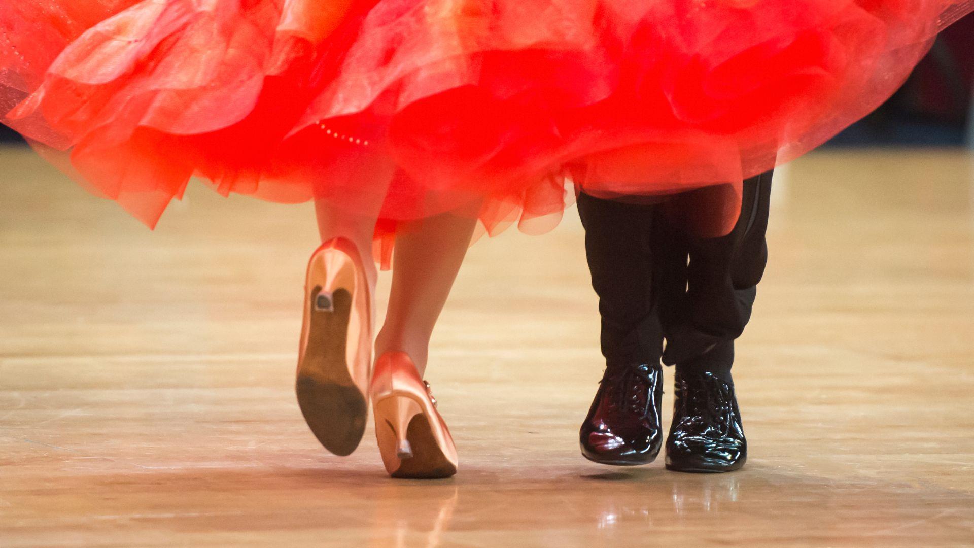Photo showing the legs and feet of two ballroom dancers. She is wearing a red dress & gold shoes. He's wearing black trousers & shoes