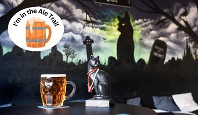 A pint of beer and a black cat set against a mural featuring a black cat and over-stamped with an I'm in the ale trail logo