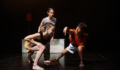 2 people crouching and communicating in dance while another stands behind observing. On a stage, dimmed lighting