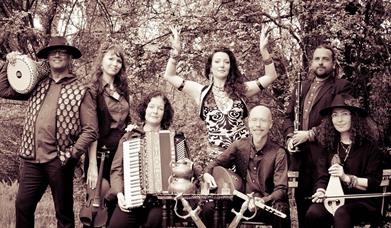 Black and white image of seven people with instruments standing in a wooded area and smiling to camera