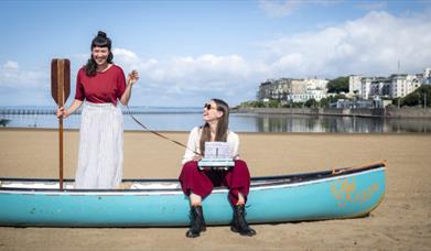 Photograph of Beth Calvery and Beth Roberts sat in a blue boat on the beach at Marine Lake.