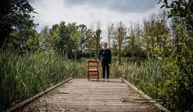 A young person stands by a chair at the end of a wooden walkway. There are trees and plants around them