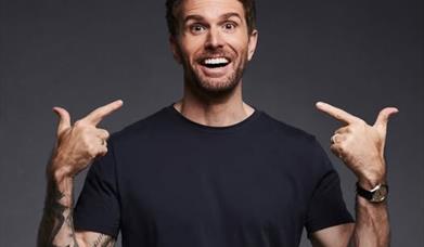 TV Presenter Joel Dommett wearing a black tee shirt is facing the camera and pointing with both hands to his smiling face