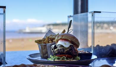 A platter of burger and chips in an open window with the view of a beach with a pier and town in the background