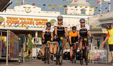 A group of four cyclists riding under the Grand Pier sign in Weston-super-Mare as they ride up the boardwalk at the finish of the Chase The Sun bike r