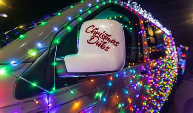 A VW van decorated in twinkling Christmas lights and the words Christmas Dubs on the wing mirror promoting the Vee Dub Christmas Cruz drives around We