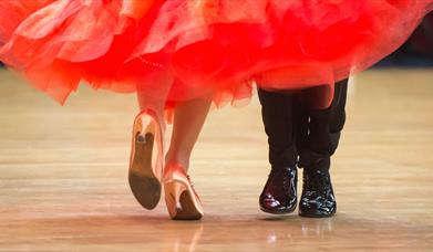 photo showing the lower legs and feet of ballroom dancers on a wooden dancefloor. She is wearing a red dress and gold shoes and he is wearing black tr