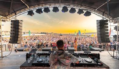 View from behind a DJ on his decks looking out across the stage onto a huge crowd at an outdoor music festival