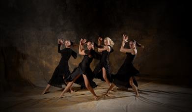 Four female dancers in flowing black dresses perform a synchronised routine on a dimly lit stage with a textured, earthy backdrop. They strike dramati