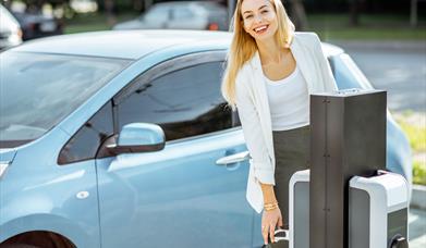 A blonde lady reaching for an EV charger to charge her blue car