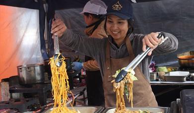 A lady stall holder at the Eat Weston Food Festival holding a tong full of noodles and another full of vegetables over two hot food platters