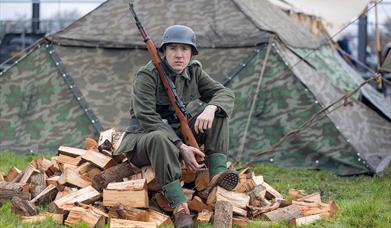 A soldier in WW2 uniform holding a rifle and sitting on a pile of split logs at the Weston-super-Mare Helicopter Museum Military History weekend