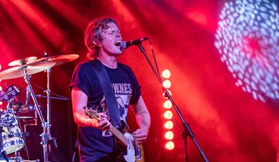 Singer Alex Lipinski playing guitar and singing into a microphone while performing a concert on stage at the Grand Pier, Weston-super-Mare