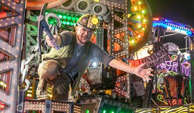A performer in a miner's helmet and with a mining pick axe on board a brightly coloured illuminated float at the Weston-super-Mare Carnival
