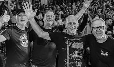 Black and white photo of four band members waving at the camera with a large crowd behind