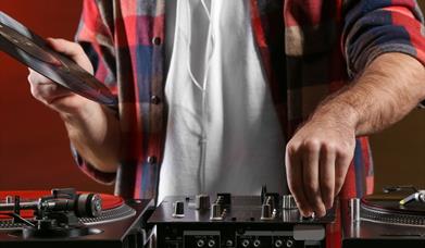 A man in a checked shirt and white undershirt (cropped without a head) holding a record above some decks