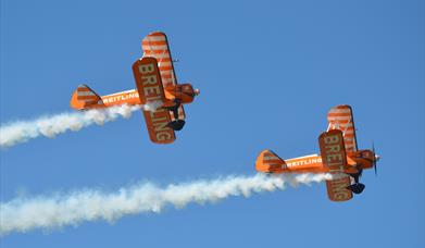 Two orange bi-planes with white plumes of smoke flying across a blue sky