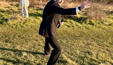 A man in a dark suit impersonating James Bond while in the background is someone in a white police crime scene suit