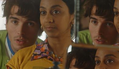 heads and shoulders of a young couple looking into mirrors so that you can see them reflected back. White male with a green jumper and and Asian femal