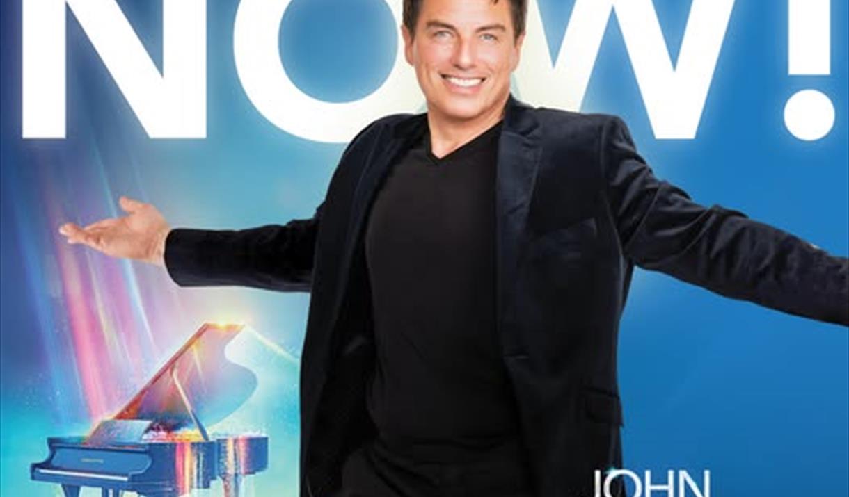 John Barrowman dressed all in black standing with his arms outstretched in front of a Grand Piano