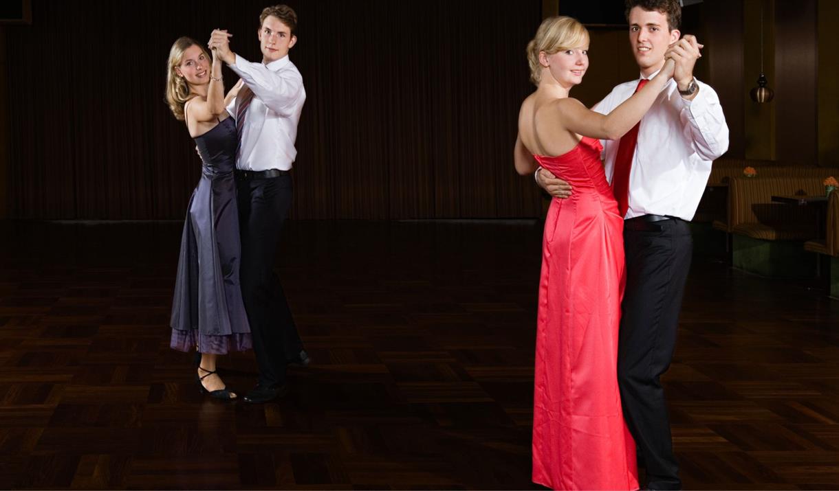 Two pairs of ballroom dancers. The men are in white shirts and black trousers and the ladies are wearing a red dress and a blue dress