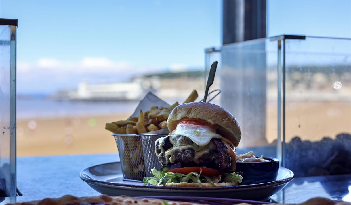 A platter of burger and chips in an open window with the view of a beach with a pier and town in the background