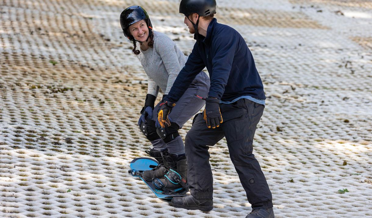 A smiling lady in a helmet on a snowboard receiving tuition from an instructor at the Mendip Activity Centre