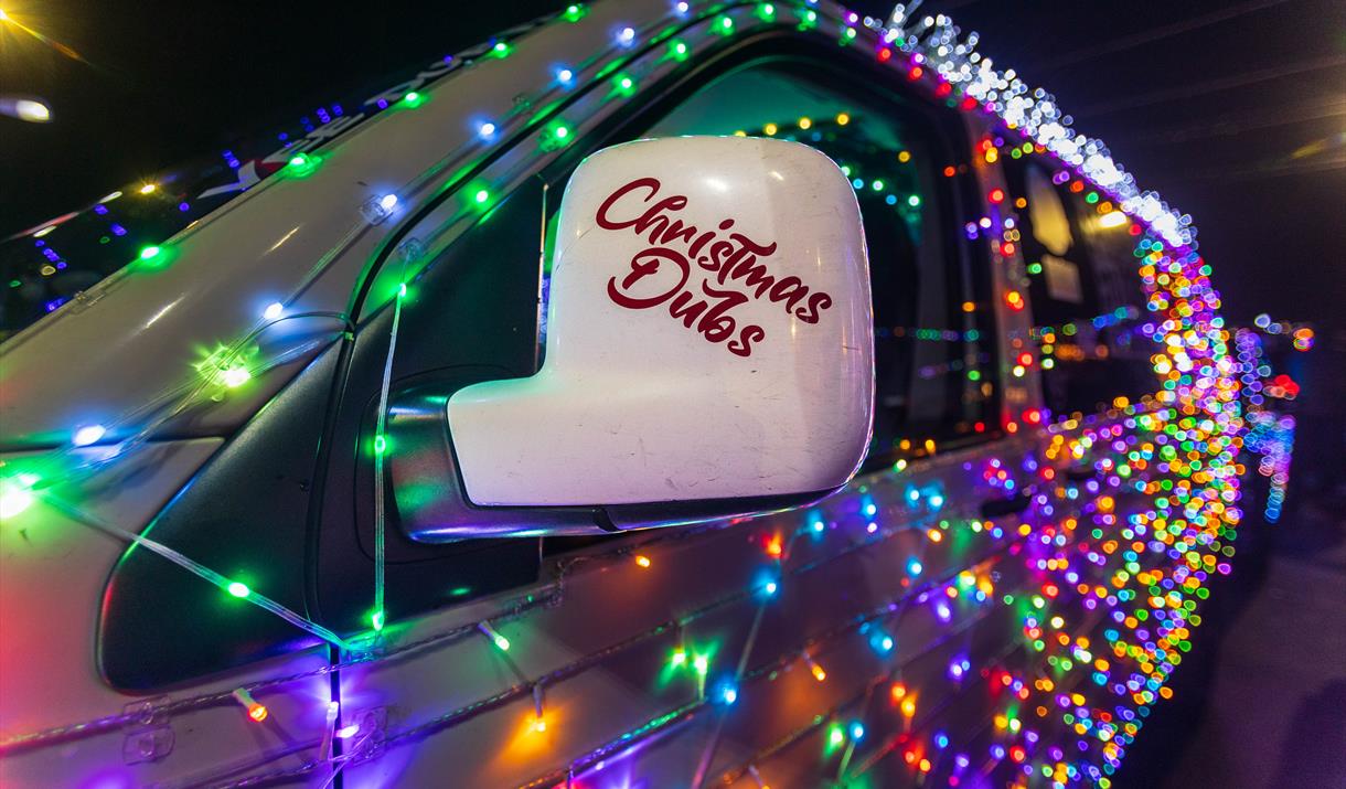 A VW van decorated in twinkling Christmas lights and the words Christmas Dubs on the wing mirror promoting the Vee Dub Christmas Cruz drives around We