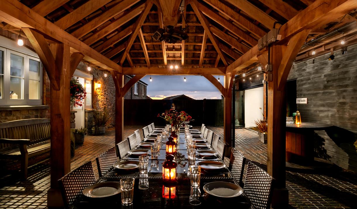 A large dining area under wooden beams which opens out to the evening sky. The large table at this Webbington Farm Holiday cottage is lit with lanters