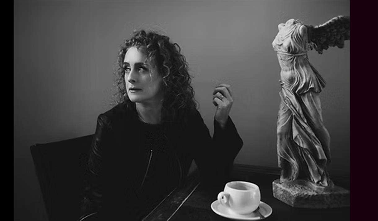 Black and white photo of singer Briana Corrigan with a white cup and saucer on a table next to a headless statue
