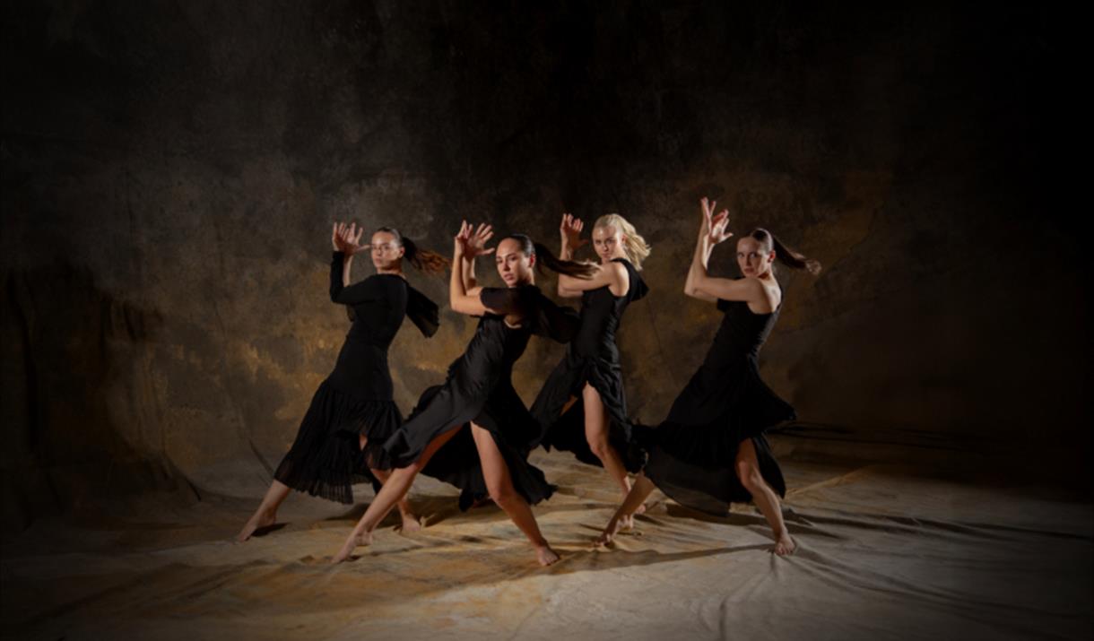 Four female dancers in flowing black dresses perform a synchronised routine on a dimly lit stage with a textured, earthy backdrop. They strike dramati